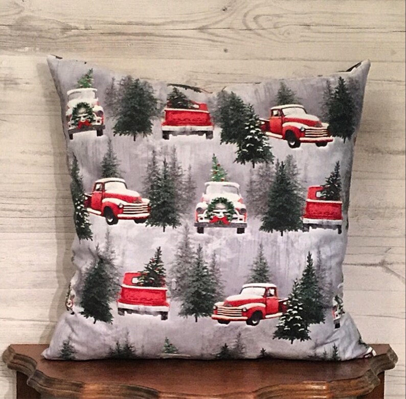 May include: A gray throw pillow with a red truck and Christmas tree pattern. The truck is carrying a Christmas tree and is driving through a snowy forest.