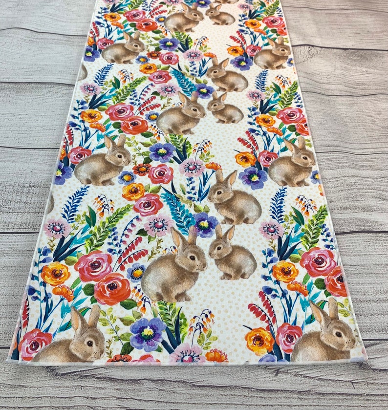 May include: A white table runner featuring a repeating pattern of brown bunnies and colourful flowers. The background is a white fabric with small white dots.