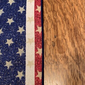 May include: A close-up of a blue and white American flag table runner with red, white, and gold glitter accents. The runner is on a wooden surface.