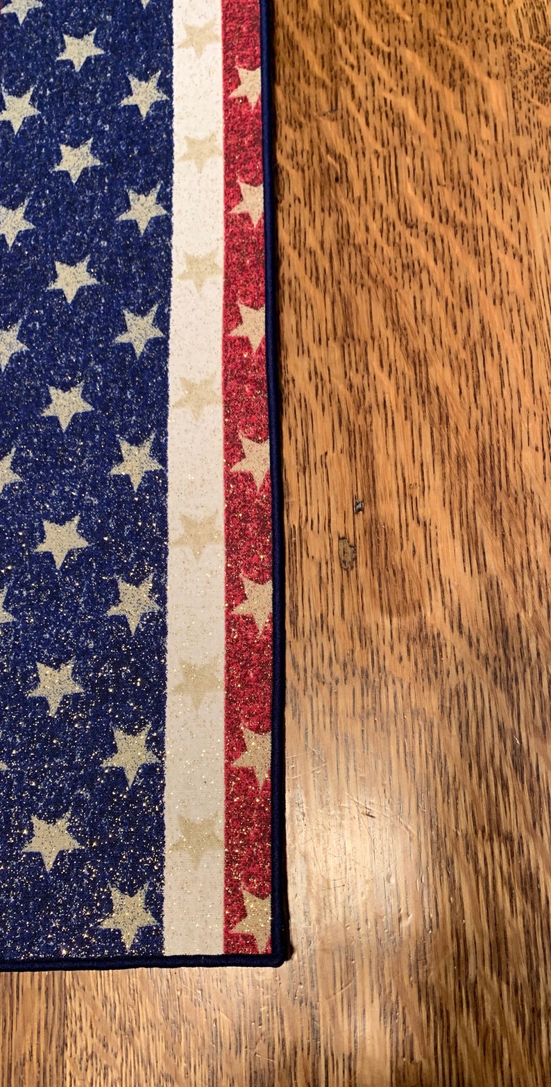 May include: A close-up of a blue and white American flag table runner with red, white, and gold glitter accents. The runner is on a wooden surface.