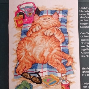 May include: Cross-stitch kit featuring a relaxed orange cat on a beach towel with the words "DO NOT DISTURB!" in pink. Includes sunglasses, a drink, and flip-flops. Finished size is 8" x 16".