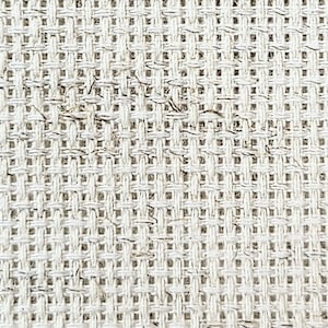 May include: Close-up of a cream-colored, woven fabric with a grid-like pattern. The fabric has a slightly rough texture and visible fibers, suggesting a natural material. The weave is tight, creating a consistent, textured surface.