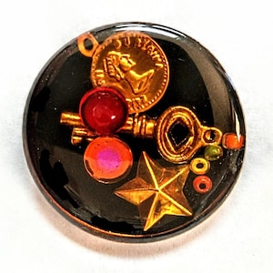 May include: A round, black resin cabochon with a gold key, a gold star, and red and pink beads. The cabochon also features a gold coin with the inscription "SU PRIM", a gold keyhole, and a gold star.