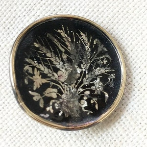 May include: A round, black button with a gold rim. The button features a silver design of a bouquet of flowers and wheat.