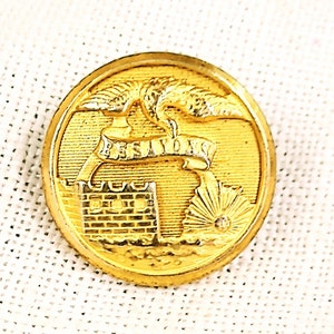 May include: A gold-colored button with a raised design. The design features a stylized image of a castle with a banner that reads "FESIVOVS" above it. The castle is surrounded by a stylized image of a sun with rays.