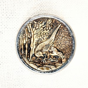May include: A round, silver-toned button with a raised, detailed design of a pheasant standing on a branch with leaves. The pheasant is gold-colored and has a detailed design.