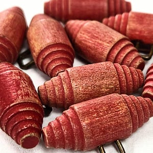 May include: A collection of red wooden beads with a grooved design and attached metal loops. The beads are arranged in a circular pattern against a white background. The beads have a rustic, vintage appearance.
