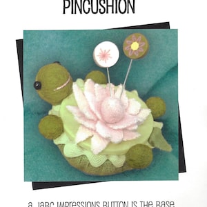 May include: A green felt turtle pincushion with a pink felt flower on top. The turtle is upside down, with the flower facing up. The pincushion is labeled "Lazy Turtle Pincushion" and "a JaBc Impressions Button is the base for this upside-down pincushion!"