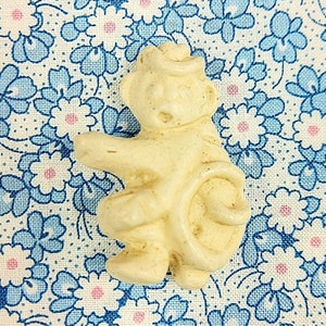 May include: A white ceramic figurine of a monkey wearing a hat. The monkey is sitting with its legs crossed and its arms raised.