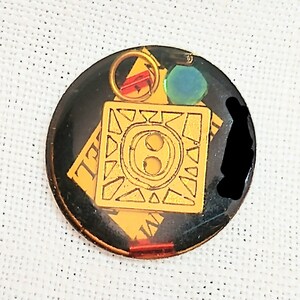 May include: A round, black and gold button with a gold design in the center. The design features a circle with two smaller circles inside, surrounded by a square with a geometric pattern. The button has a red and green accent.