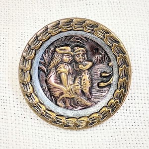 May include: A vintage brass button with a raised design of two children standing in a field. The button has a gold-colored rim with a decorative border.