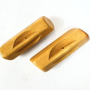 May include: Two vintage, rectangular wooden drawer pulls in a light brown color. Each pull features a carved leaf-like design and a small hole for mounting. The pulls are displayed on a white background.
