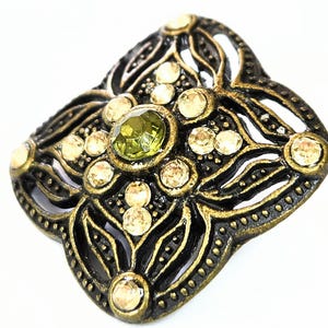 May include: Ornate, antique-style button with a black and gold finish. The button features a central green gemstone surrounded by smaller, clear gemstones. The design is symmetrical with a floral motif, perfect for embellishing garments or crafts.