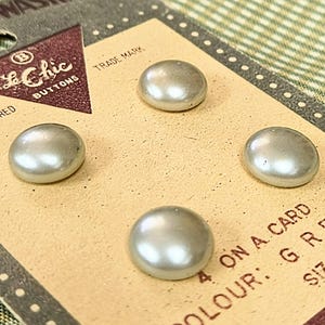 May include: Four pearlescent gray buttons on a vintage card. The card has the text "Le Chic Buttons" and "Trade Mark". The buttons are oval-shaped and have a glossy finish. The card also has the text "4 on a card" and "Color: Grey".