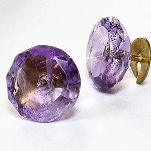 May include: Two faceted, purple amethyst cufflinks with gold-colored metal backings. The gemstones have a brilliant cut, showcasing their internal clarity and color variations. The cufflinks are set against a white background.