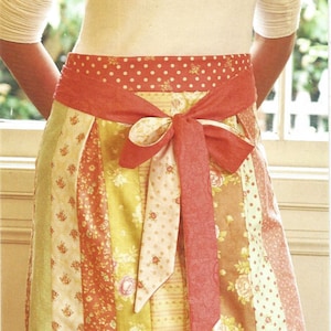 May include: A patchwork apron with a red polka dot trim and bow. The apron features floral and dotted patterns in shades of yellow, orange, and pink. Text on the apron reads "a wraparound jelly roll apron".