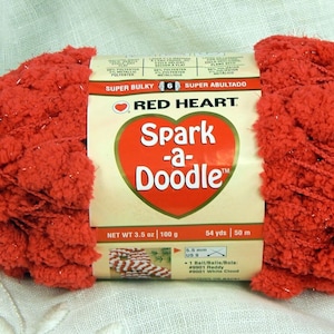 May include: A skein of Red Heart Spark-a-Doodle yarn in a bright red color with a sparkly finish. The yarn is super bulky and has a pom-pom texture. The label reads "Red Heart Spark-a-Doodle" and "Super Bulky".