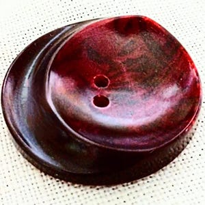 May include: A large, swirled button with two holes. The button is a deep burgundy color with darker marbling throughout. The button has a glossy finish and a slightly irregular shape, suggesting it is handmade.