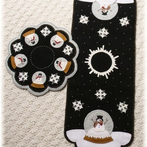 May include: Two black felt table runners with white snowflake designs and snowmen. One runner is round and features snowmen in snow globes. The other runner is rectangular and features a snowman in a snow globe and a sun.