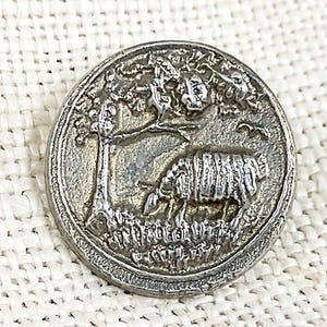 May include: A silver metal button with a raised design of a sheep standing under a tree. The button has a circular shape and a raised rim.