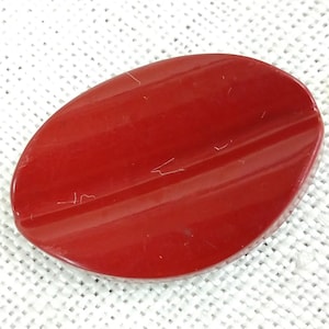 May include: A glossy, oval-shaped, red button with a slightly curved surface. The button has a smooth finish and a few minor scratches. The button is likely made of plastic or a similar material. It could be used for clothing or crafts.