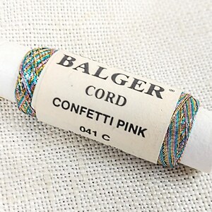 May include: A spool of Balger Cord Confetti Pink thread, with a metallic finish in shades of teal, gold, pink, and silver. The spool is labeled "Balger Cord Confetti Pink 041 C".