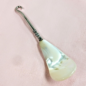 May include: A shoe horn with a pearlescent white scoop and a silver-colored metal handle. The handle has a curved hook at the top and a decorative band near the scoop. The shoe horn is set against a pink background.