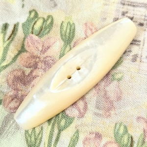 May include: An elongated, diamond-shaped button with a smooth, off-white, pearlescent finish. The button features a central ridge running lengthwise and two small, circular holes positioned in the middle for attachment to fabric or garments.