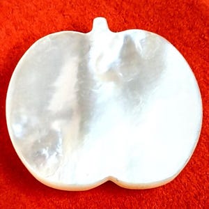 May include: A pearly white, apple-shaped decorative item. The surface has a shimmering, iridescent quality. The apple shape has a stem at the top and a slight indentation at the bottom. The background is a solid red.