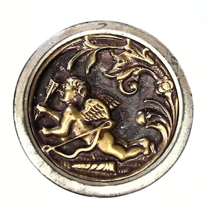 May include: A round, antique-style button with a silver-colored rim. The center features a gold-colored cherub with a bow and arrow, set against a dark brown background. Ornate gold-colored floral designs surround the cherub.