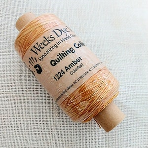 May include: A spool of Weeks Dye Quilting Collection thread in the color 1224 Amber. The spool is brown with a label that says "Weeks Dye", "Specializing in Hand Dyed", "Quilting Collection", "1224 Amber", "Colorfast", and "Mechanical Blvd. Garner, NC 27529 USA 8/7.OVERDE".