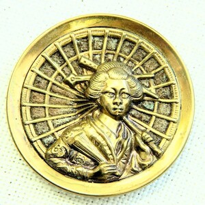 May include: A gold-toned metal button with a raised image of a woman in a traditional Japanese kimono holding a fan. The background is a stylized sunburst design.