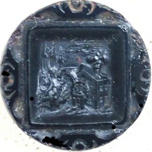 May include: A dark, circular object with a square, raised central design. The square features a detailed relief scene, possibly depicting a building and figures. The outer rim has an ornate, swirling pattern. The object appears to be made of metal.