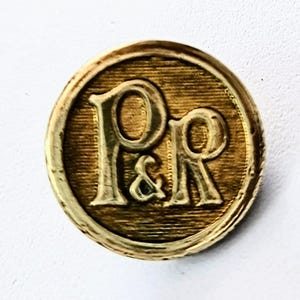 May include: A round, gold-colored button with the letters "P&R" embossed in a serif font. The button has a textured surface and a raised edge, giving it a vintage or antique appearance. The background is a plain white.