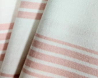 May include: Close-up of a folded white fabric with horizontal stripes in a soft pink hue. The fabric appears to be soft and textured, with the stripes adding a simple, yet elegant design. The folds create shadows and depth.