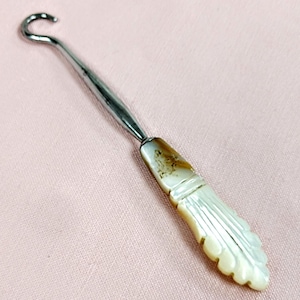 May include: A vintage button hook with a silver-toned metal shaft and a curved hook. The handle is made of a cream-colored material with a carved leaf design and brown accents. The hook is designed to assist with buttoning shoes and clothing.