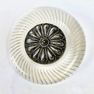 May include: A circular button featuring a clear, transparent outer rim with etched radial lines. The center is an inset metallic silver-toned disc with a raised, embossed floral pattern consisting of multiple petals and a small gold-toned dot.
