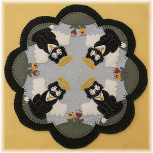 May include: A round, multi-colored felt rug with a black, white, and gray design of four cats with halos and wings. The cats are arranged in a circular pattern around a central point. The rug has a scalloped edge.