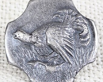 May include: A silver-toned metal button with a detailed relief of a pheasant in a grassy field. The button has a unique, four-lobed shape and a textured, aged appearance, suggesting a vintage or antique style.