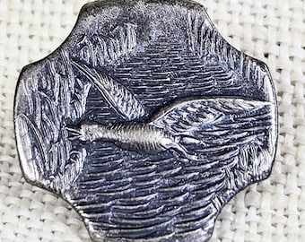 May include: A silver-toned metal pin with a detailed relief of a bird in flight over water, surrounded by reeds and a mountainous background. The pin has a unique, four-lobed shape and a vintage aesthetic.