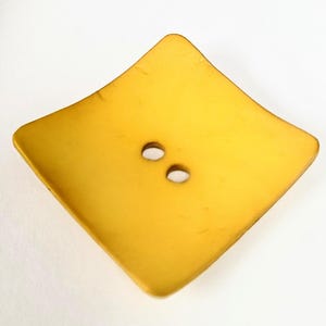 May include: A large, golden-yellow, diamond-shaped button with two small, round holes. The button has a slightly textured surface and rounded edges, suggesting a vintage or handcrafted aesthetic. Ideal for sewing or craft projects.