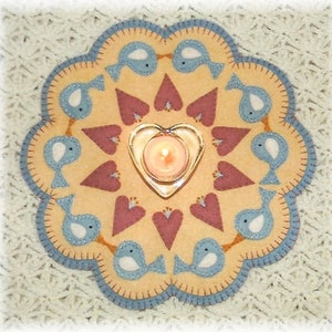 May include: A circular felt table runner with a blue and brown border. The center features a design of hearts and birds with a candle in a heart-shaped holder.