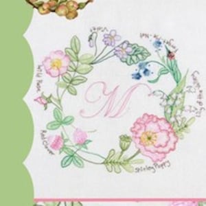 May include: A white embroidery pattern with a floral wreath design. The wreath is made up of various flowers and leaves, including wild roses, clover, forget-me-nots, and a Shirley poppy. The center of the wreath features the letter "M" in pink. The text "Crabapple Hill" and "Hedgerow Wreath" are also included in the design.