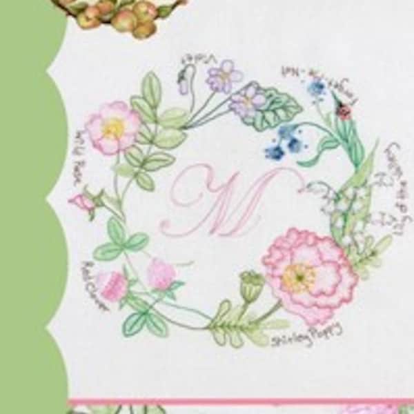 May include: A white embroidery pattern with a floral wreath design. The wreath is made up of various flowers and leaves, including wild roses, clover, forget-me-nots, and a Shirley poppy. The center of the wreath features the letter "M" in pink. The text "Crabapple Hill" and "Hedgerow Wreath" are also included in the design.