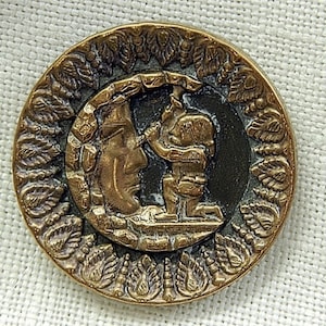 May include: A round, antique-style button with a detailed relief design. The button features a central scene of a figure kneeling before a crescent moon, surrounded by a decorative border of leaves. The button has a bronze finish.