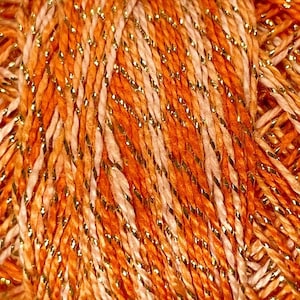 May include: Close-up of a ball of yarn with a mix of orange, peach, and cream-colored strands. The yarn is interwoven with strands of gold metallic thread, creating a shimmering effect. Ideal for knitting and crochet projects.