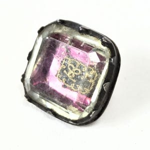 May include: A square button featuring a faceted, translucent purple center stone encased in a dark, metallic frame. The center of the purple stone displays a small, intricate gold-colored emblem with a decorative border, set against a dark background.