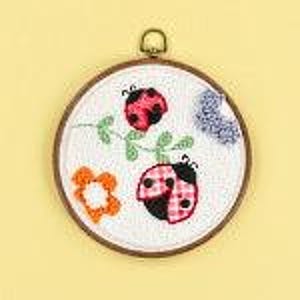 May include: Embroidered hoop art featuring two red and black ladybugs with white polka dots on a white background. The ladybugs are on a green vine with green leaves. A small orange flower is in the lower left corner.