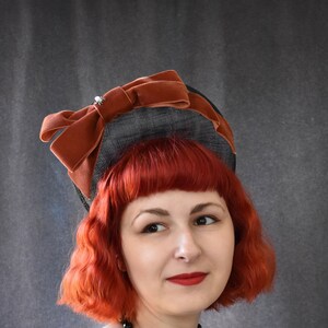 May include: A woman with red hair wearing a black and brown hat with a velvet bow and a black beaded necklace. She is wearing a black and orange gingham dress with white lace trim.
