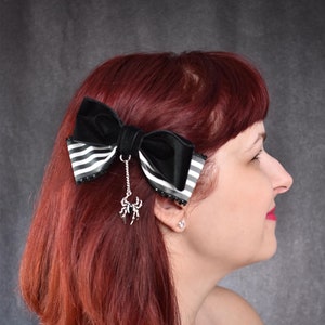 May include: A black and white striped bow with a black velvet bow on top. The bow has a silver spider charm hanging from it. The bow is worn in a woman's red hair.